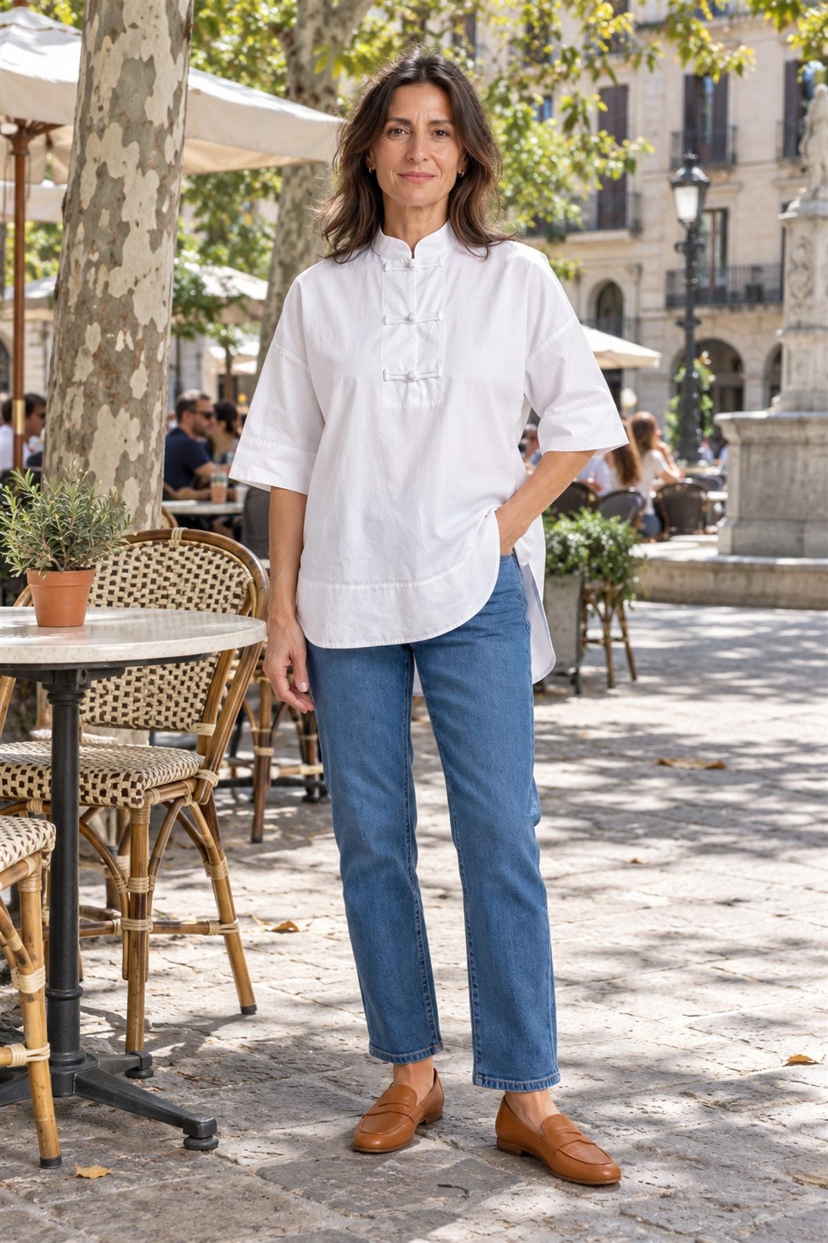 Olivia, camisa blanca por fuera del vaquero, en la terraza de una plaza con árboles